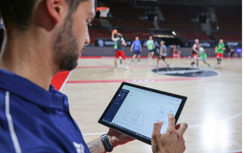 Ilustrasi pelatih NBA sedang menganalisis data statistik permainan menggunakan tablet di pinggir lapangan.
